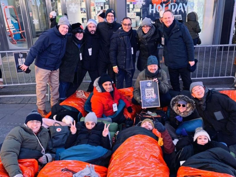 Group of participants bundled in winter gear and orange sleeping bags on a sidewalk in Times Square for the World's Big Sleep Out